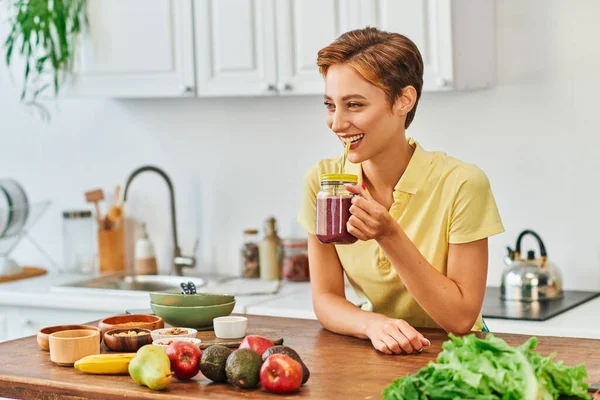 Mulher vegetariana sorrindo bebendo delicioso smoothie de pote de pedreiro com palha na cozinha — Fotografia de Stock