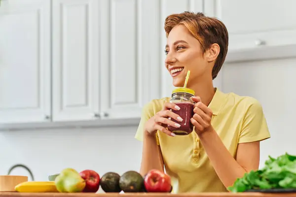 Mulher vegetariana alegre segurando smoothie fresco no frasco de pedreiro com palha e olhando para longe na cozinha — Fotografia de Stock