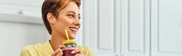 Mulher alegre com smoothie vegetariano em frasco de pedreiro com palha olhando para longe na cozinha, banner — Fotografia de Stock