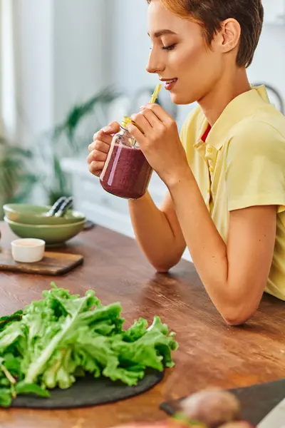 Молодая вегетарианка держит банку масона с вкусным смузи возле свежего салата на кухонном столе — стоковое фото
