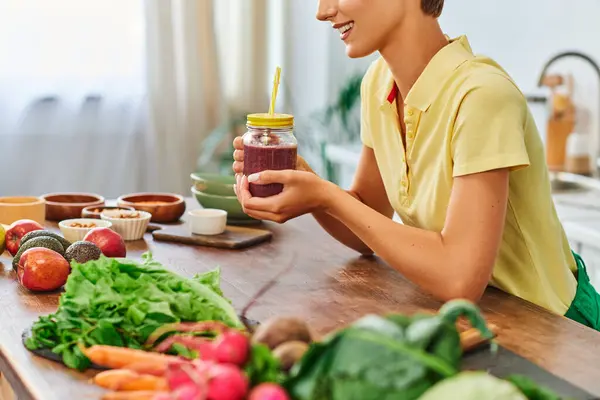 Donna ritagliata con frullato vegetariano in barattolo di muratore vicino a ingredienti a base vegetale sul tavolo della cucina — Foto stock