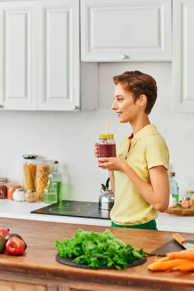 Vista laterale di donna vegetariana sorridente in possesso di vaso di muratore con frullato fresco in cucina moderna — Foto stock