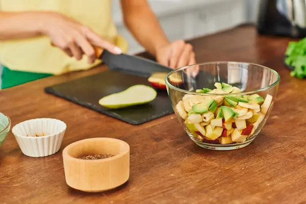Schüssel mit gehackten Früchten nahe zugeschnittener Frau, die Birne auf unscharfem Hintergrund schneidet, vegetarischer Salat — Stockfoto
