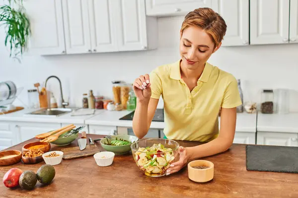 Усміхнена жінка приправляє свіжий фруктовий салат з насінням кунжуту на кухні, смачна вегетаріанська дієта — стокове фото