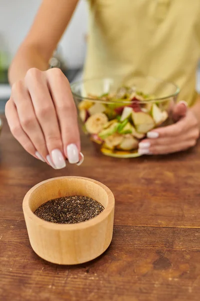 Обрезанная вегетарианка берет семена кунжута из деревянной чаши во время приготовления фруктового салата дома — стоковое фото
