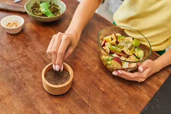 Обрізаний вид жінки, що приймає насіння кунжуту з дерев'яної миски біля свіжого фруктового салату, вегетаріанська дієта — стокове фото