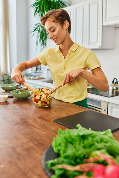 Mujer joven y alegre mezclando ensalada de frutas frescas mientras cocina comida vegetariana en la cocina moderna - foto de stock