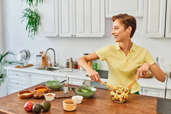 Cheerful woman mixing vegetarian salad bear fresh fruits and looking away in modern kitchen — Stock Photo