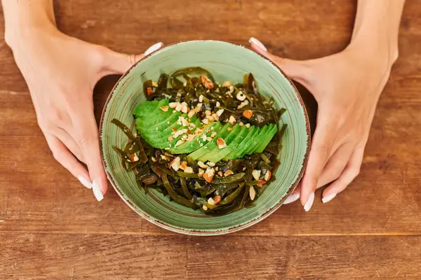 Vista superior de manos femeninas cerca de ensalada con algas marinas y aguacate en rodajas con nueces, receta vegetariana - foto de stock
