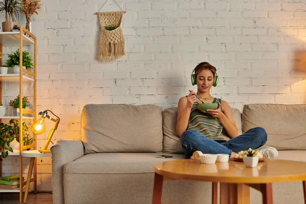 Mujer encantada en auriculares teniendo cena vegetariana en el sofá en acogedora sala de estar, noche tranquila — Stock Photo