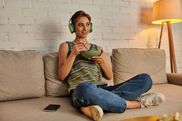 Smiling woman in headphones with salad bowl on couch in living room, vegetarian evening snack — Stock Photo