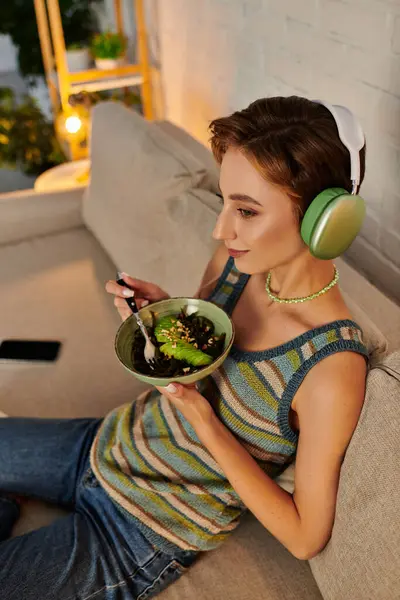 Мрійлива вегетаріанка в навушниках тримає салат з водоростями і нарізаним авокадо на дивані вдома — стокове фото
