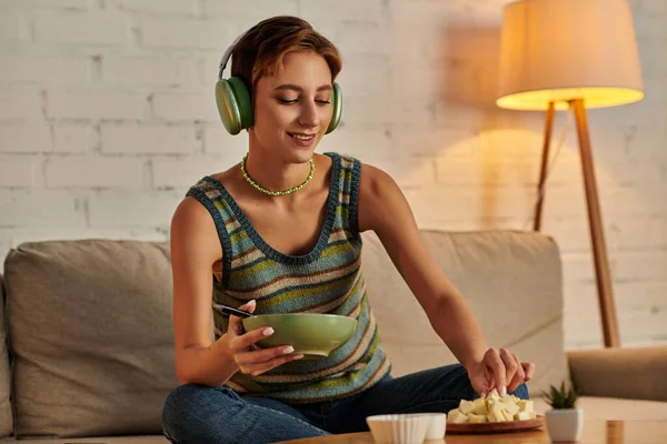 Smiley woman in headphones holding salad bowl and taking tofu cheese, vegetarian dinner on couch — Stock Photo