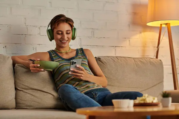 Happy veggie woman in headphones networking on smartphone while sitting with salad bowl on couch — Stock Photo
