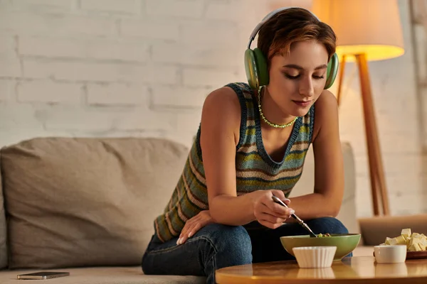 Junge Frau mit Kopfhörer isst vegetarischen Salat in der Nähe von Tofu-Käse, während sie abends auf dem Sofa sitzt — Stockfoto