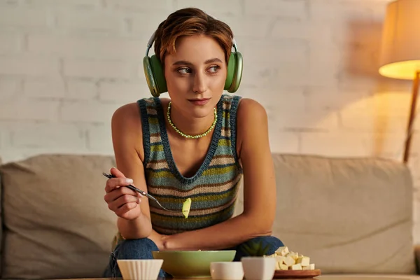 Mujer de ensueño en auriculares mirando hacia otro lado mientras toma merienda vegetariana por la noche en el sofá en casa - foto de stock