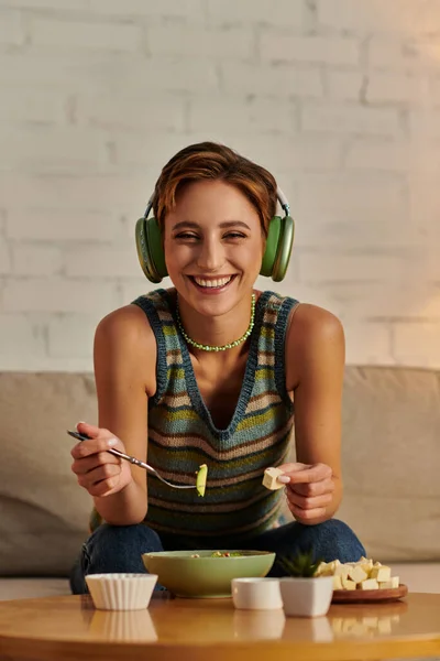 Mujer vegetariana alegre en auriculares mirando a la cámara cerca de ensalada a base de plantas y queso de tofu — Stock Photo