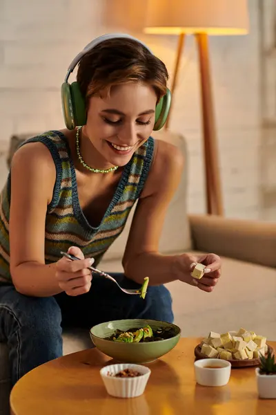 Mulher alegre em fones de ouvido jantar com salada vegetariana e queijo tofu no sofá em casa — Fotografia de Stock