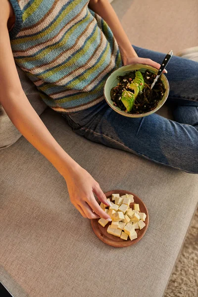 Vista ritagliata della donna con insalata vegetariana seduta sul divano vicino al formaggio tofu, spuntino serale — Foto stock