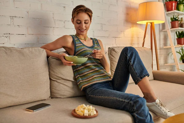 Donna felice seduta sul divano vicino al tofu formaggio e smartphone e mangiare insalata vegetariana — Foto stock