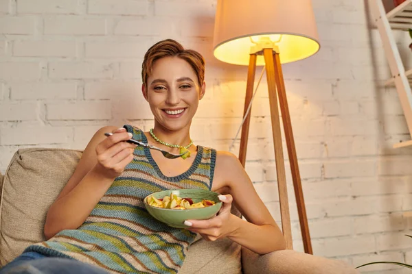 Alegre joven mujer mirando a la cámara mientras se sienta con ensalada vegetariana en acogedor salón - foto de stock