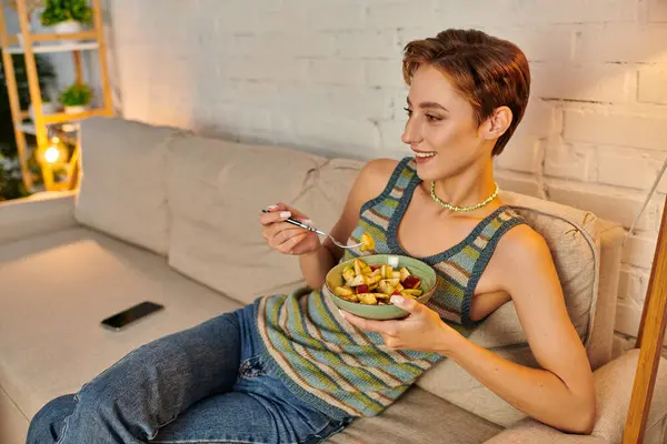 Улыбающаяся женщина сидит с миской вкусного вегетарианского фруктового салата на диване в гостиной — стоковое фото