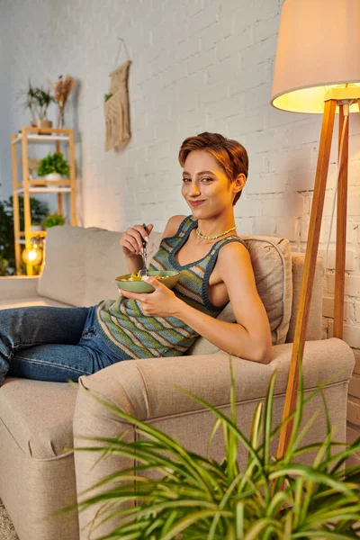 Jovem mulher vegetariana sorrindo para a câmera enquanto come salada de frutas no sofá na acolhedora sala de estar — Fotografia de Stock
