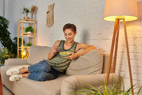 Delighted veggie woman sitting on couch near lamp and eating fresh fruit salad on cozy sofa — Stock Photo