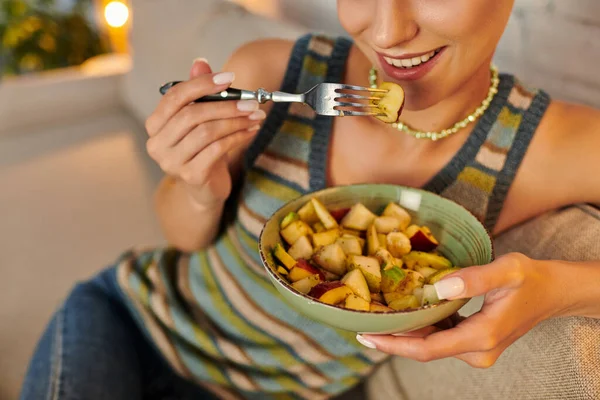 Vista cortada de mulher vegetariana comendo deliciosa salada de frutas para o jantar no sofá na sala de estar — Fotografia de Stock