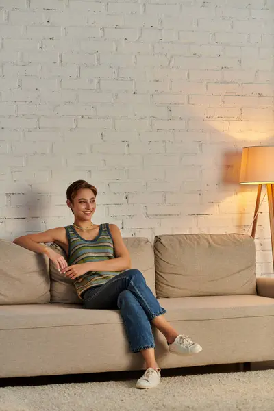 Happy young woman sitting on cozy couch near lamp in modern living room, leisure in quiet evening — Stock Photo
