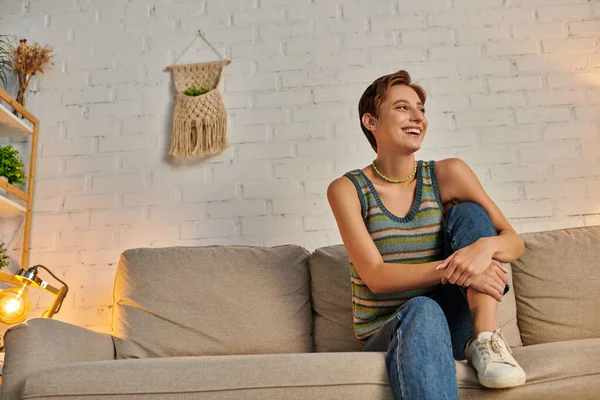 Joven mujer emocionada mirando hacia otro lado en el sofá acogedor en la sala de estar moderna, feliz vida en casa - foto de stock