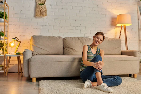 Happy young woman sitting on floor near couch in modern living room and smiling at camera in evening — Stock Photo