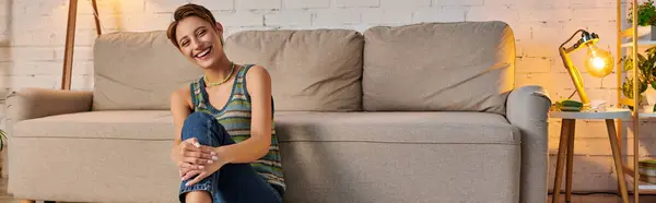 Joyous young woman sitting near couch in cozy living room and looking at camera, horizontal banner — Stock Photo