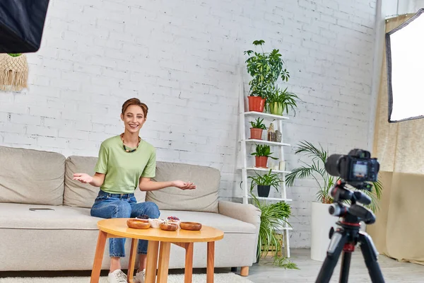 Улыбающаяся женщина представляет вегетарианскую еду во время видео блога о растительных диетах в зеленой гостиной — стоковое фото