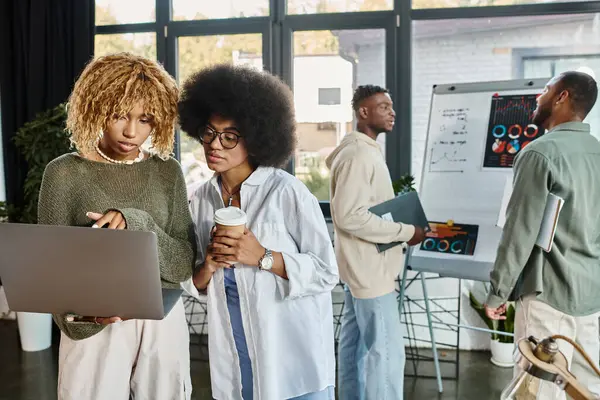 Zwei Frauen schauen auf Laptop mit Kaffeetasse in der Hand mit ihren männlichen Freunden auf Hintergrund, Geschäft — Stockfoto