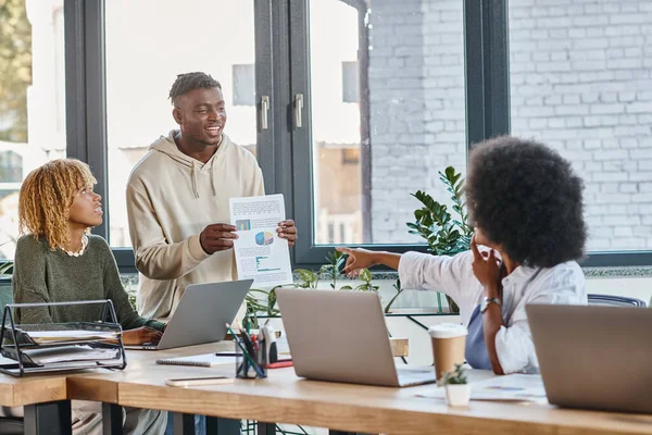 Good looking african american man showing to his female friends graphics, pointing finger, coworking — Stock Photo