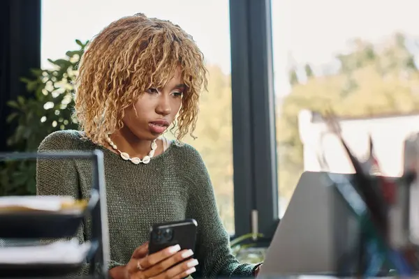 Femme attrayante avec les cheveux bouclés regardant l'ordinateur portable et tenant le téléphone mobile, processus de travail — Photo de stock