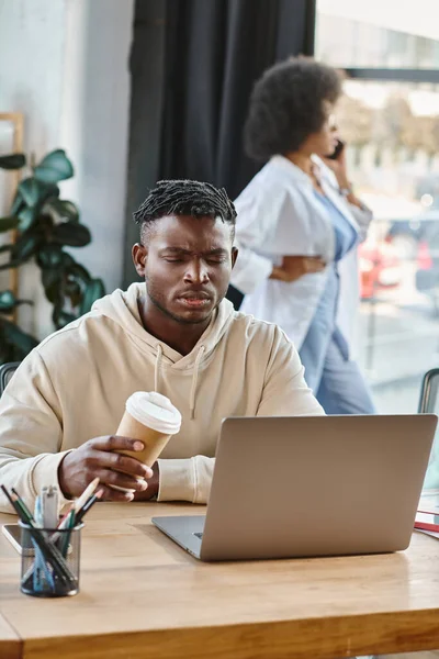 Сфокусированный красивый мужчина смотрит на ноутбук, держа свой кофе с другом на заднем плане, коворкинг — стоковое фото