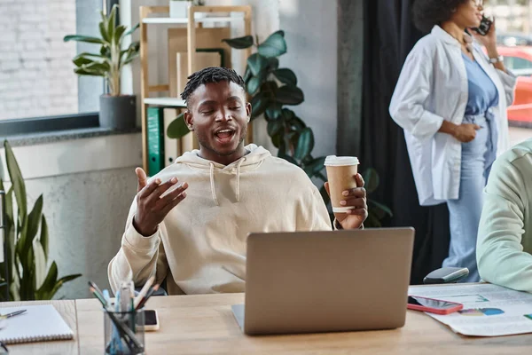 Fröhlicher junger Mann gestikuliert und schaut auf Laptop mit seiner Freundin im Hintergrund, Coworking — Stockfoto
