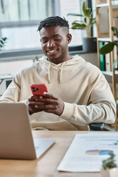 Homem americano africano alegre olhando alegremente para seu telefone na frente do laptop, processo de trabalho — Fotografia de Stock
