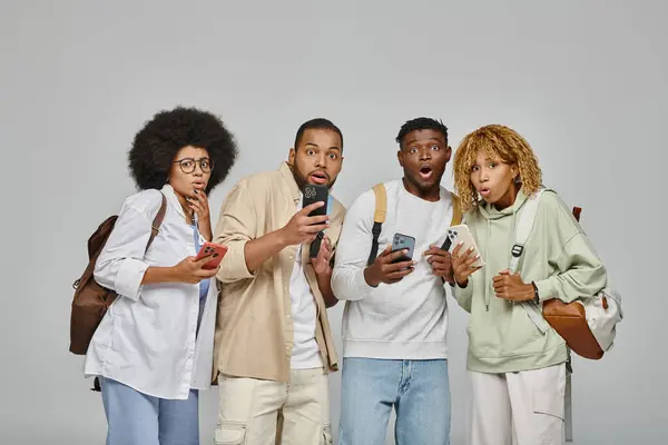 Schockierte afrikanisch-amerikanische Freunde mit Rucksäcken mit Handys, die überrascht in die Kamera schauen, Studenten — Stockfoto