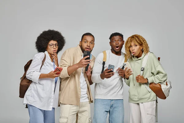 Sorprendió a los amigos afroamericanos con mochilas con teléfonos mirando sorprendidos a la cámara, estudiantes - foto de stock