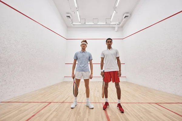 Young interracial male friends in sportswear looking at camera and holding racquets for squash — Stock Photo