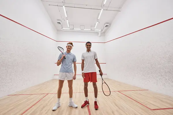 Feliz interracial macho amigos en sportswear mirando cámara y sosteniendo raquetas para squash - foto de stock