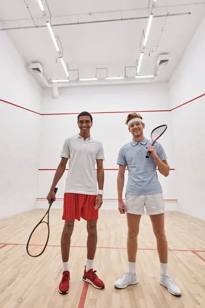 Vertical shot, happy interracial male friends looking at camera and holding racquets for squash — Stock Photo