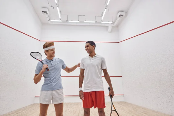 Feliz interracial masculino amigos em sportswear sorrindo e segurando raquetes para squash dentro do tribunal — Fotografia de Stock