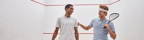 Happy interracial men  in sportswear smiling and holding racquets for squash inside of court, banner — Stock Photo