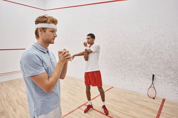 Young redhead man with headband warming up near african american friend inside squash court — Stock Photo