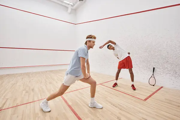 Junger rothaariger Mann mit Stirnband, der in der Nähe eines afrikanisch-amerikanischen Freundes im Squash-Court Ausfallbewegungen macht — Stockfoto