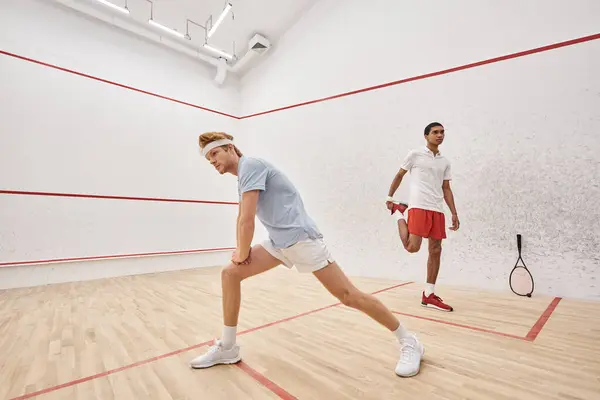 Sporty redhead man with headband doing lunges near african american friend inside squash court — Stock Photo